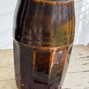 May include: A vintage brown stoneware crock with a dark interior. The crock has a cylindrical shape with a slightly flared top and a series of vertical ridges. The exterior has a glossy finish with variations in color, from dark brown to a lighter, amber hue.
