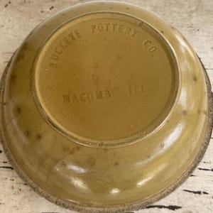 May include: A vintage, round, stoneware bowl with a light brown glaze. The bottom of the bowl features the embossed text "BUCKEYE POTTERY CO. MACOMB ILL.". The rim has a darker, unglazed band.