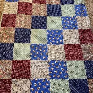 May include: A patchwork quilt featuring a grid of square fabric panels in various patterns and colors, including deep red, navy blue, light blue, and floral prints. The quilt is constructed with visible seams between each individual square.