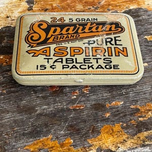 May include: Vintage Spartan Brand aspirin tin. The rectangular tin is beige with orange and black lettering. The text reads "24 5 Grain Spartan Brand Pure Aspirin Tablets 15¢ Package". The tin has a distressed appearance.
