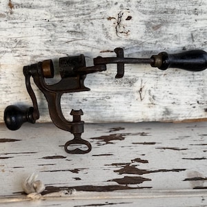 May include: An antique, cast iron corkscrew with a black wooden handle and a crank. The corkscrew is mounted on a base with a clamp and a screw. The metal has a dark, aged patina, suggesting its vintage.