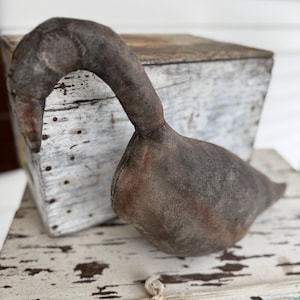 May include: A handcrafted, stuffed goose figurine with a curved neck and a dark, weathered appearance. The goose is primarily gray with brown accents, suggesting an aged or vintage aesthetic. The goose is placed on a distressed, white-painted surface.