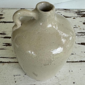 May include: A vintage, off-white stoneware jug with a rounded body, a handle, and a narrow opening. The jug has a glossy finish and appears to be handmade. The surface shows slight imperfections, adding to its rustic charm. This antique piece is suitable for home decor.
