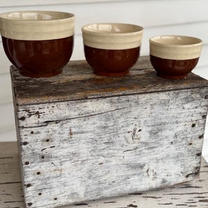 May include: Three ceramic bowls in varying sizes, each with a two-tone design. The bowls have a cream-colored upper section and a rich brown lower section. They are arranged on a weathered, white-painted wooden surface.