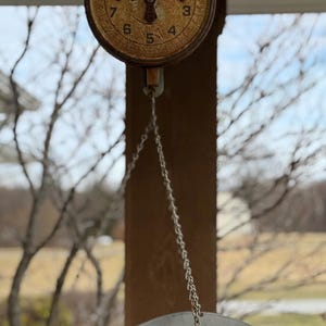 May include: An antique hanging scale with a round, aged face and a metal pan. The scale's face has black numbers and a needle pointing to zero. A chain suspends the pan below.