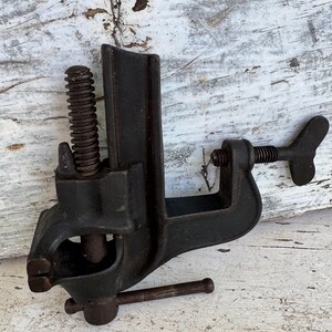 May include: A dark gray cast iron bench vise featuring a vertical threaded screw mechanism, a horizontal tightening handle, and a side-mounted wing nut screw for securing the tool to a surface. The metal body has a textured, weathered finish.