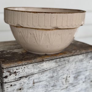 May include: A beige ceramic mixing bowl with a ribbed rim and a decorative pattern on the lower half. The bowl is sitting on a weathered wooden surface, suggesting a vintage or rustic aesthetic.