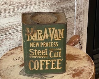 May include: An antique green metal coffee tin with the text "KARAVAN NEW PROCESS Steel Cut COFFEE" in gold lettering. The tin is rectangular with a lid and is placed on a round, distressed wooden surface.