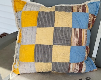 May include: A patchwork pillow with squares of yellow, blue, brown, and gray fabric. The pillow is sitting on a white surface.