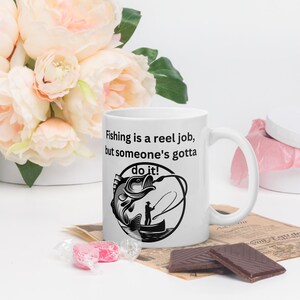Op de afbeelding: Een witte keramische koffiemok met een zwart-witte illustratie van een visser die een vis binnenhaalt. De tekst op de mok luidt "Fishing is a reel job, but someone's gotta do it!"