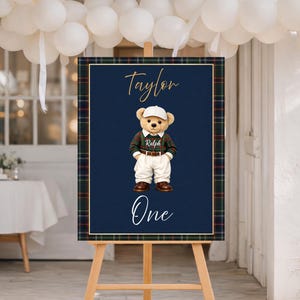 Puede incluir: Un letrero azul marino con un borde a cuadros presenta un oso de peluche con gorra, suéter y pantalones. El letrero dice "Taylor" y "One" en escritura cursiva. El letrero se muestra en un caballete de madera.