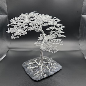 May include: A handcrafted silver wire tree sculpture. The tree features a detailed wire trunk, branches, and leaves, and is mounted on a dark, textured base. The sculpture is set against a black background.