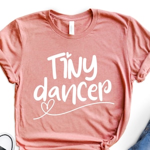 May include: A pink t-shirt with the text "Tiny dancer" in white lettering. The shirt is on a white background with a pair of black sneakers, a pair of denim shorts, and a coffee mug.