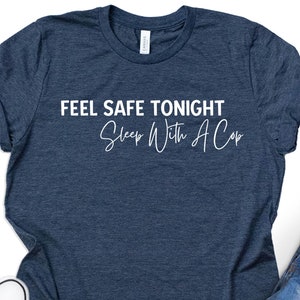 May include: A navy blue t-shirt with the text "FEEL SAFE TONIGHT Sleep With A Cop" in white script. The shirt is paired with denim shorts and black sneakers. A cup of coffee sits nearby.