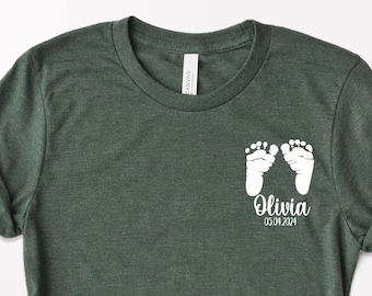 May include: A dark green t-shirt with a white design of two baby footprints and the text "Olivia 03.04.2024".