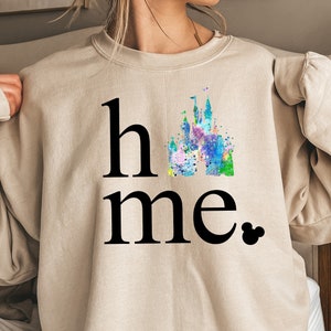 Puede incluir: Una sudadera beige con las palabras "home" y una ilustraci&oacute;n de acuarela de un castillo. El castillo est&aacute; en tonos de azul, verde, rosa y morado. La palabra "home" est&aacute; en negro y el castillo est&aacute; en el centro de la palabra.