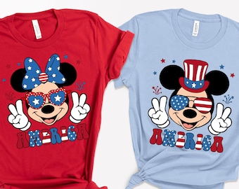 May include: Red and light blue t-shirts featuring Mickey and Minnie Mouse with patriotic designs. Mickey wears a top hat and sunglasses with the American flag, while Minnie has a bow and sunglasses. The word "AMERICA" is printed on both shirts.