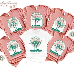 May include: Five t-shirts with a retro design featuring the text "Palm Springs" and palm trees. The text "Before the Rings" is printed below the design. Four of the shirts are a light pink color and one is white.