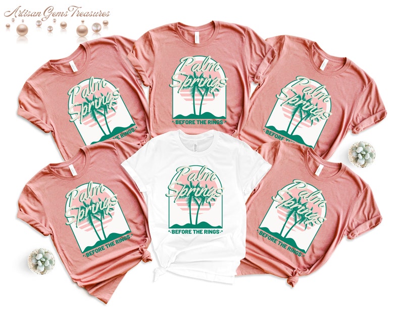 May include: Five t-shirts with a retro design featuring the text "Palm Springs" and palm trees. The text "Before the Rings" is printed below the design. Four of the shirts are a light pink color and one is white.