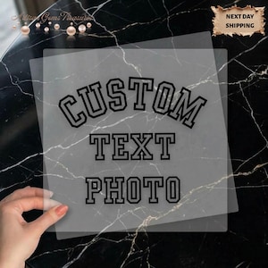 May include: A transparent sheet with the words "CUSTOM TEXT PHOTO" in a bold, black, collegiate-style font. The sheet is held over a black and white marble surface. The words are centered on the sheet.
