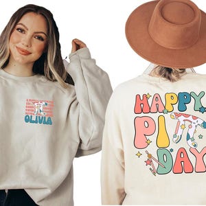 May include: A light beige sweatshirt with a graphic design on the front that says "A Pi-day a day to celebrate... Olivia" and a graphic design on the back that says "Happy Pi Day" with a colorful pie and stars.