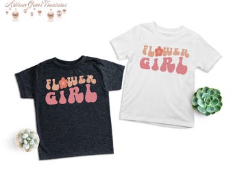 May include: Two t-shirts with the text "Flower Girl" in a retro font with a flower graphic. One shirt is dark grey and the other is white.