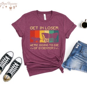 Op de afbeelding: Een maroon t-shirt met een vintage-stijl grafische afbeelding met de tekst "Get in loser, we're going to die of dysentery". De afbeelding toont een wagen en een persoon op de achtergrond.