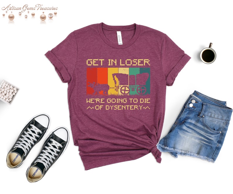 Op de afbeelding: Een maroon t-shirt met een vintage-stijl grafische afbeelding met de tekst "Get in loser, we're going to die of dysentery". De afbeelding toont een wagen en een persoon op de achtergrond.