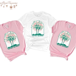 May include: Three t-shirts with a graphic design of palm trees and the text "Palm Springs - Before the Rings". The shirts are in pink and white.