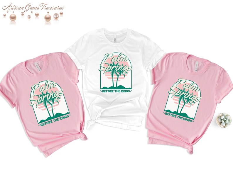 May include: Three t-shirts with a graphic design of palm trees and the text "Palm Springs - Before the Rings". The shirts are in pink and white.