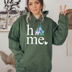 Puede incluir: Una sudadera con capucha verde con el texto "home" y una ilustraci&oacute;n colorida del castillo de Disney. El texto est&aacute; en blanco y hay un peque&ntilde;o icono de Mickey Mouse al lado de la palabra "home".