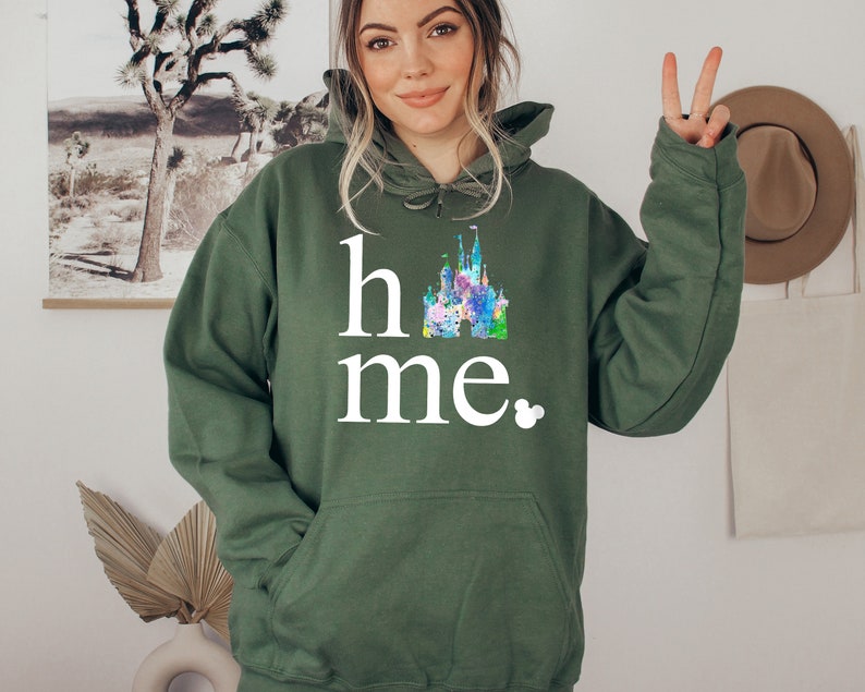 Puede incluir: Una sudadera con capucha verde con el texto "home" y una ilustraci&oacute;n colorida del castillo de Disney. El texto est&aacute; en blanco y hay un peque&ntilde;o icono de Mickey Mouse al lado de la palabra "home".