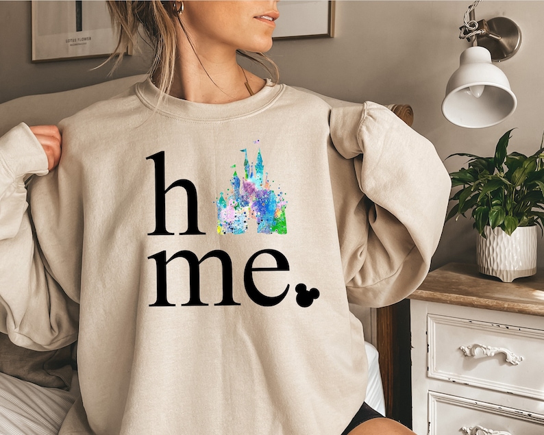Puede incluir: Una sudadera beige con las palabras "home" y una ilustraci&oacute;n de acuarela de un castillo. El castillo est&aacute; en tonos de azul, verde, rosa y morado. La palabra "home" est&aacute; en negro y el castillo est&aacute; en el centro de la palabra.