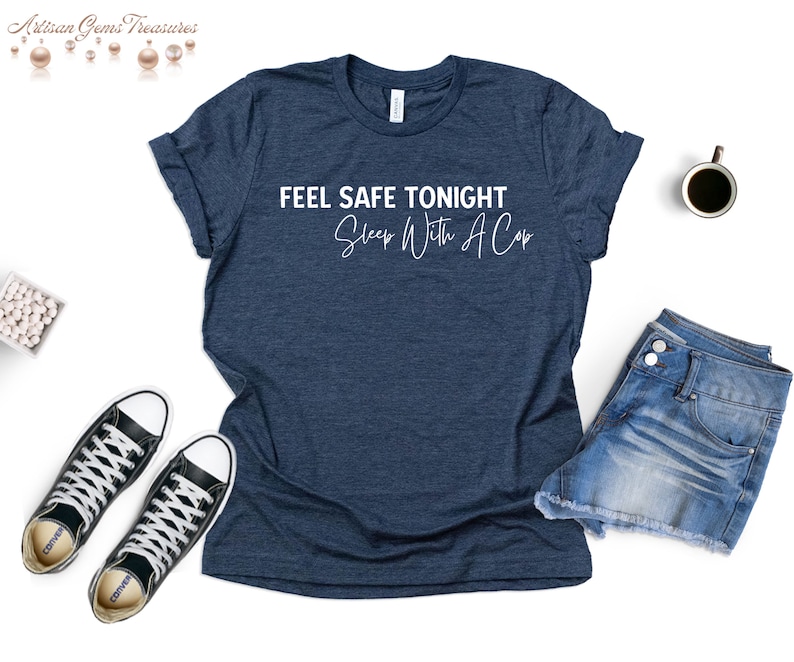 May include: A navy blue t-shirt with the text "FEEL SAFE TONIGHT Sleep With A Cop" in white script. The shirt is paired with denim shorts and black sneakers. A cup of coffee sits nearby.