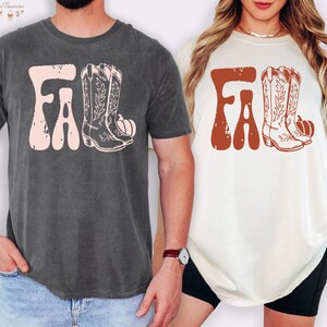 May include: Two t-shirts, one charcoal gray and one cream, each featuring a fall-themed design. The design includes the word "FALL" in a retro font, cowboy boots, and a pumpkin. The charcoal shirt has a pink design, while the cream shirt has a rust-colored design.
