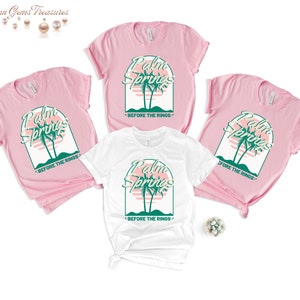 May include: Three t-shirts, two pink and one white, with a graphic design of palm trees and a sunset. The text "Palm Springs" is written in a retro font above the palm trees. The text "Before the Rings" is written below the palm trees.