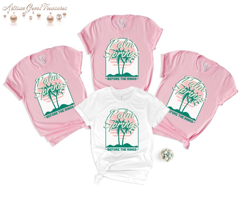 May include: Three t-shirts, two pink and one white, with a graphic design of palm trees and a sunset. The text "Palm Springs" is written in a retro font above the palm trees. The text "Before the Rings" is written below the palm trees.