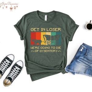 Op de afbeelding: Groen T-shirt met een vintage-stijl grafische afbeelding van een wagen en de tekst "GET IN LOSER. WE'RE GOING TO DIE ~OF DYSENTERY~"