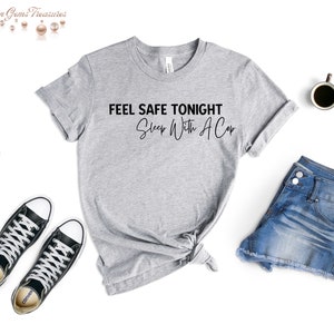 May include: A heather gray t-shirt with the text "FEEL SAFE TONIGHT Sleep With A Cop" in black. The shirt is styled with denim shorts and black sneakers. A coffee cup is also visible.