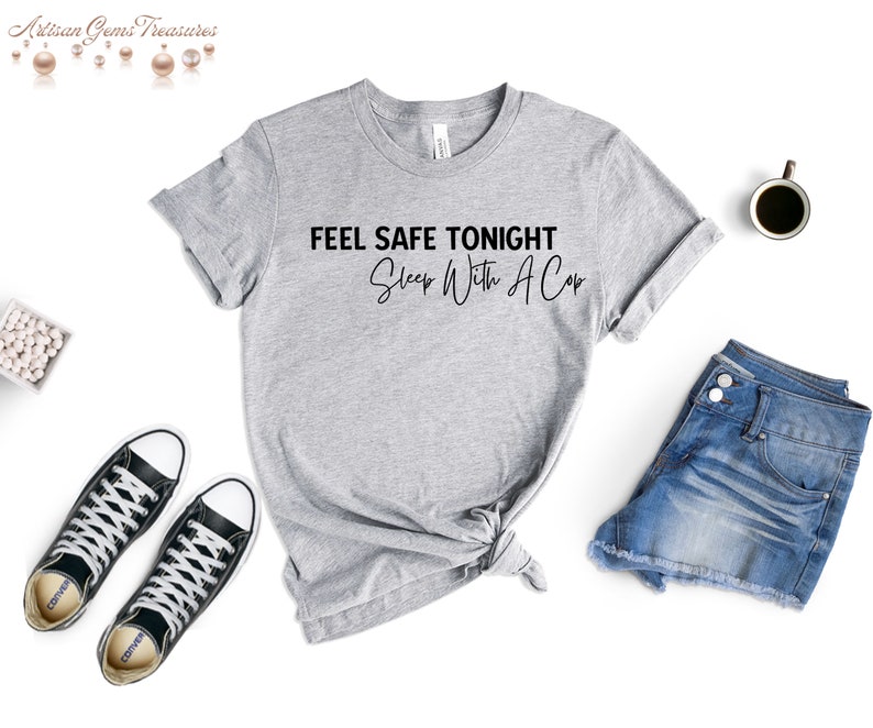 May include: A heather gray t-shirt with the text "FEEL SAFE TONIGHT Sleep With A Cop" in black. The shirt is styled with denim shorts and black sneakers. A coffee cup is also visible.