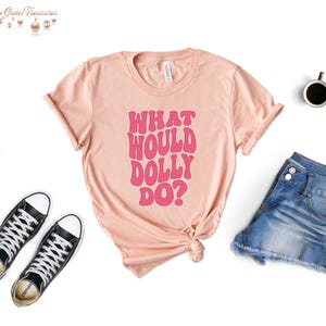 May include: A peach-colored t-shirt with the text "What Would Dolly Do?" in pink lettering. The text is in a retro font style.