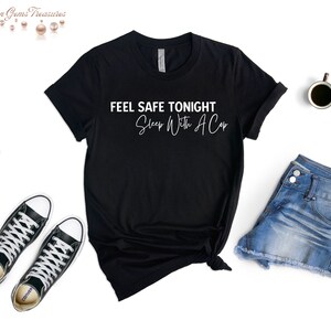 May include: Black t-shirt with the text "FEEL SAFE TONIGHT Sleep With A Cap" in white. The shirt is styled with denim shorts, black sneakers, and a cup of coffee on a white background.