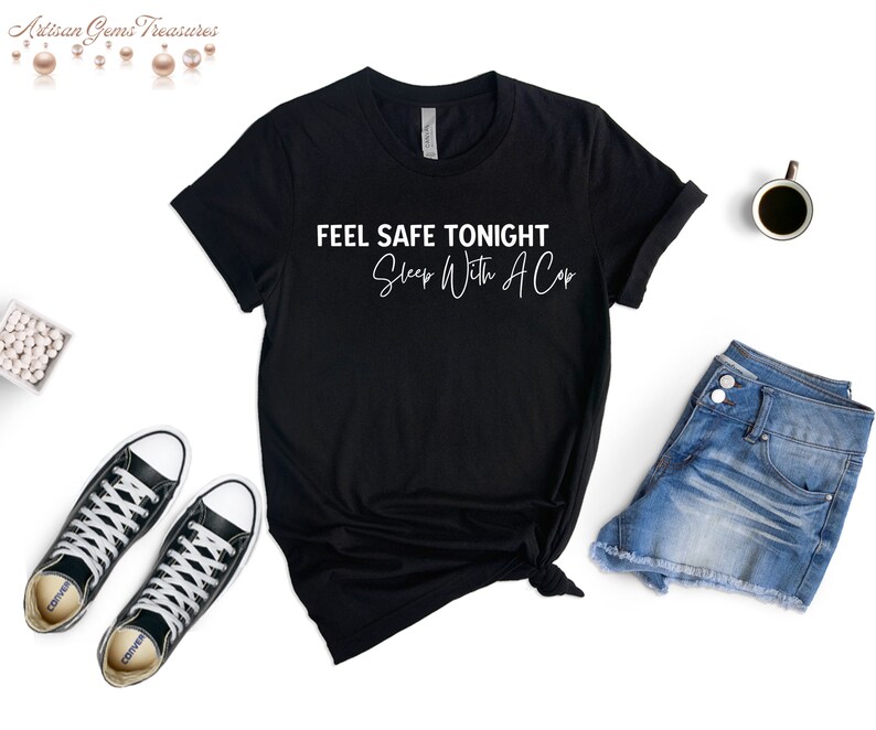 May include: Black t-shirt with the text "FEEL SAFE TONIGHT Sleep With A Cap" in white. The shirt is styled with denim shorts, black sneakers, and a cup of coffee on a white background.
