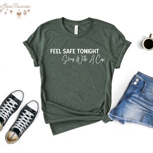 May include: A dark green t-shirt with the text "FEEL SAFE TONIGHT Sleep With A Cop" in white. The shirt is styled with denim shorts and black and white sneakers. A cup of coffee is also visible.