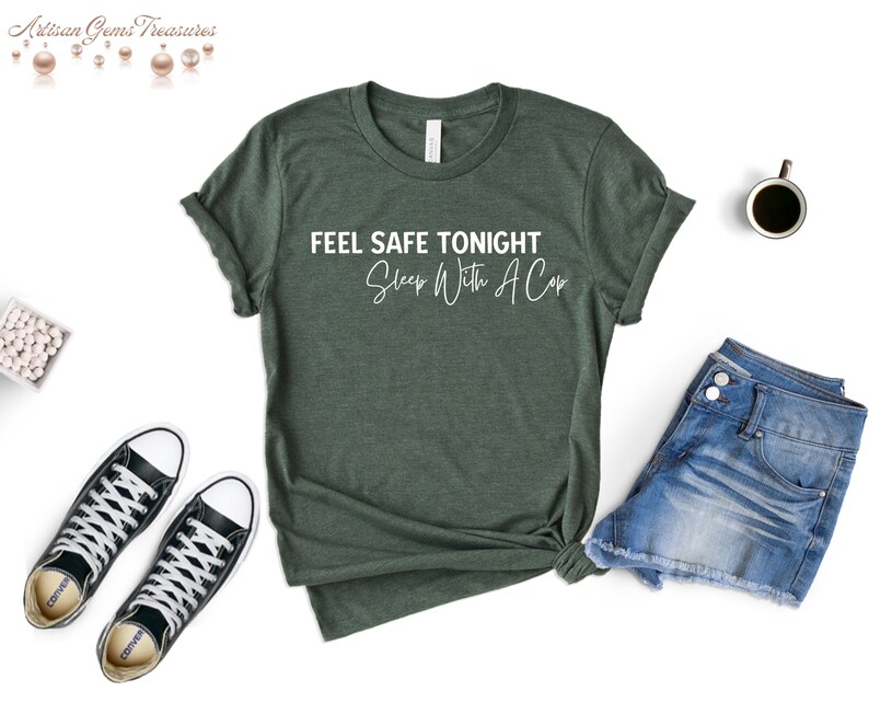 May include: A dark green t-shirt with the text "FEEL SAFE TONIGHT Sleep With A Cop" in white. The shirt is styled with denim shorts and black and white sneakers. A cup of coffee is also visible.