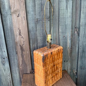 May include: A rectangular table lamp base featuring a woven basket-weave texture in a warm brown finish. The lamp includes a brass-toned metal socket, a tall brass harp, and a small black finial at the top, designed for a standard light bulb.