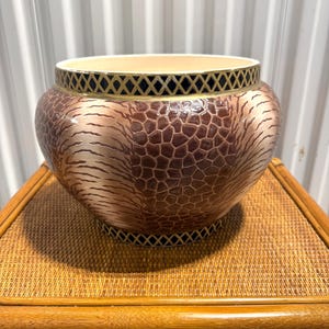 May include: A round ceramic bowl featuring a textured brown and metallic bronze finish with a reptilian scale pattern. The rim and base are decorated with a black and gold lattice design, while the interior of the bowl is a smooth, solid cream color.