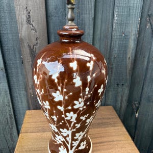 May include: A glossy brown ceramic table lamp base featuring a white painted floral branch pattern. The lamp has a bulbous body that tapers toward the base and a metallic gold-toned socket fixture at the top, resting on a circular white base.