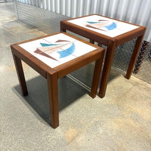 May include: Two square side tables with wooden frames and white tile tops. Each tabletop features a four-tile inlay displaying an abstract geometric design in shades of blue, brown, and tan. The tables have four straight legs and a natural wood finish.
