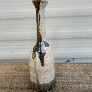 May include: A tall, slender ceramic vase with a flared rim and a narrow neck. The body features a textured, off-white speckled glaze with dark brown, vertical drip patterns running from the rim down to the base, which is finished in a dark olive green.